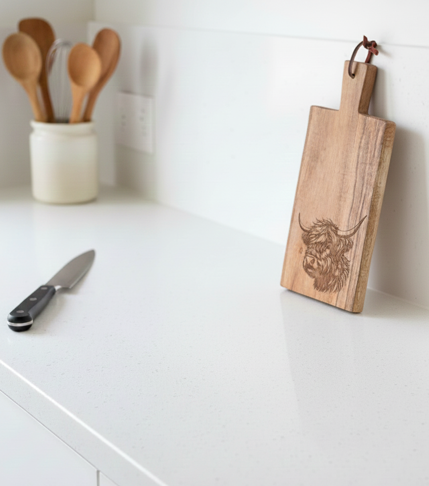 Rectangular wooden chopping board with a coo engraving, leaning against a smooth white wall on a speckled white counter.