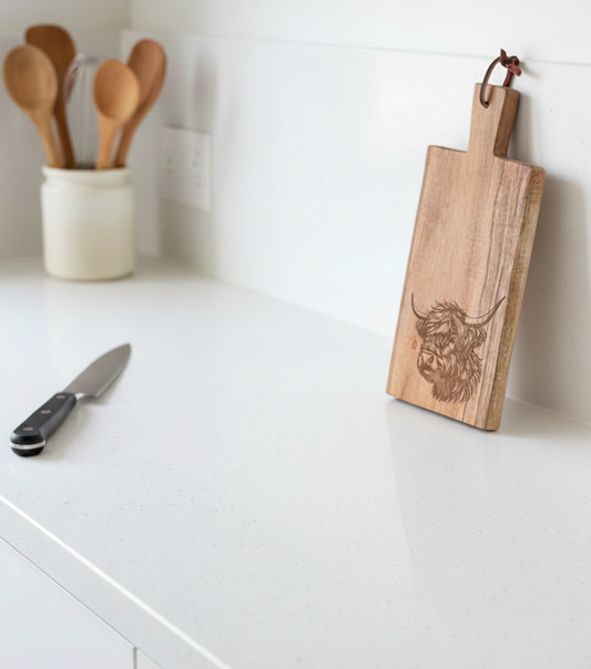 Rectangular wooden chopping board with a coo engraving, leaning against a smooth white wall on a speckled white counter.