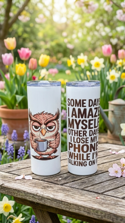 Two white tumblers on a weathered wooden table. One shows a brown owl; the other, brown text. Background of colourful blooms.