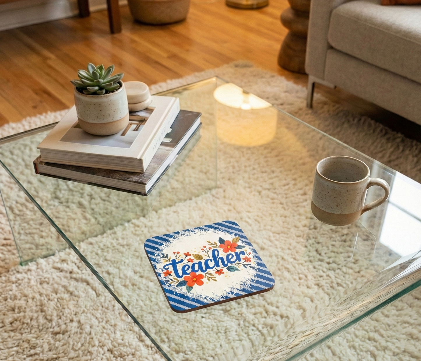 Blue striped coaster with 'teacher' in cursive and floral accents on a glass table over a white shaggy rug.