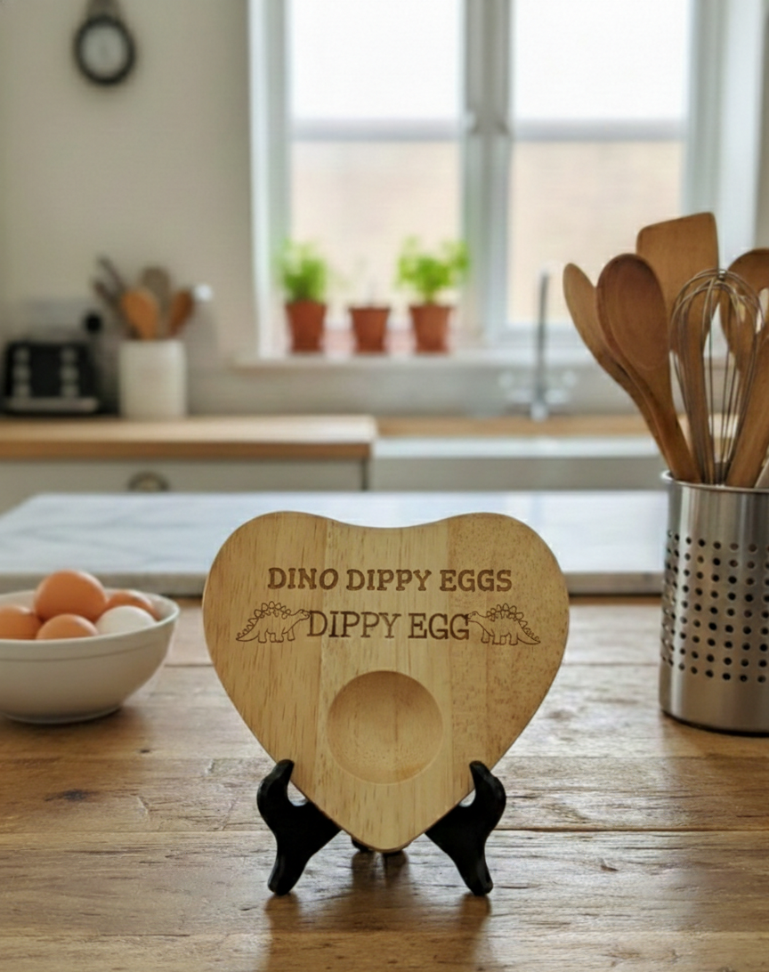 Light wooden, heart-shaped dippy egg board, engraved with text and dinosaurs, standing on a black holder.