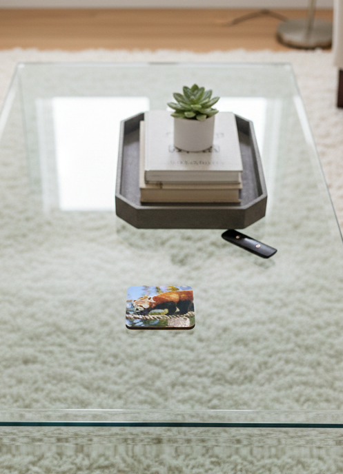 A square red panda coaster on a glass coffee table with a grey tray holding a book and green succulent in the back.