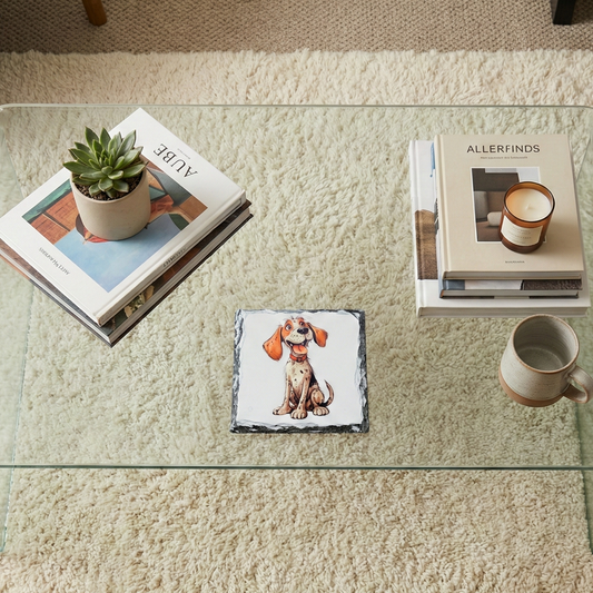 Square slate coaster with a cartoon spaniel with ginger ears, on a glass table over a cream shaggy rug.