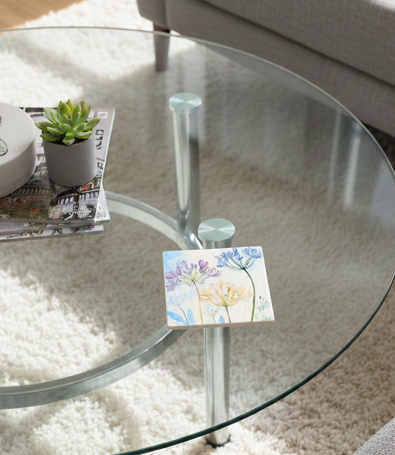 Square coaster featuring purple and yellow wildflower watercolour art on a clear glass table over a textured white rug.