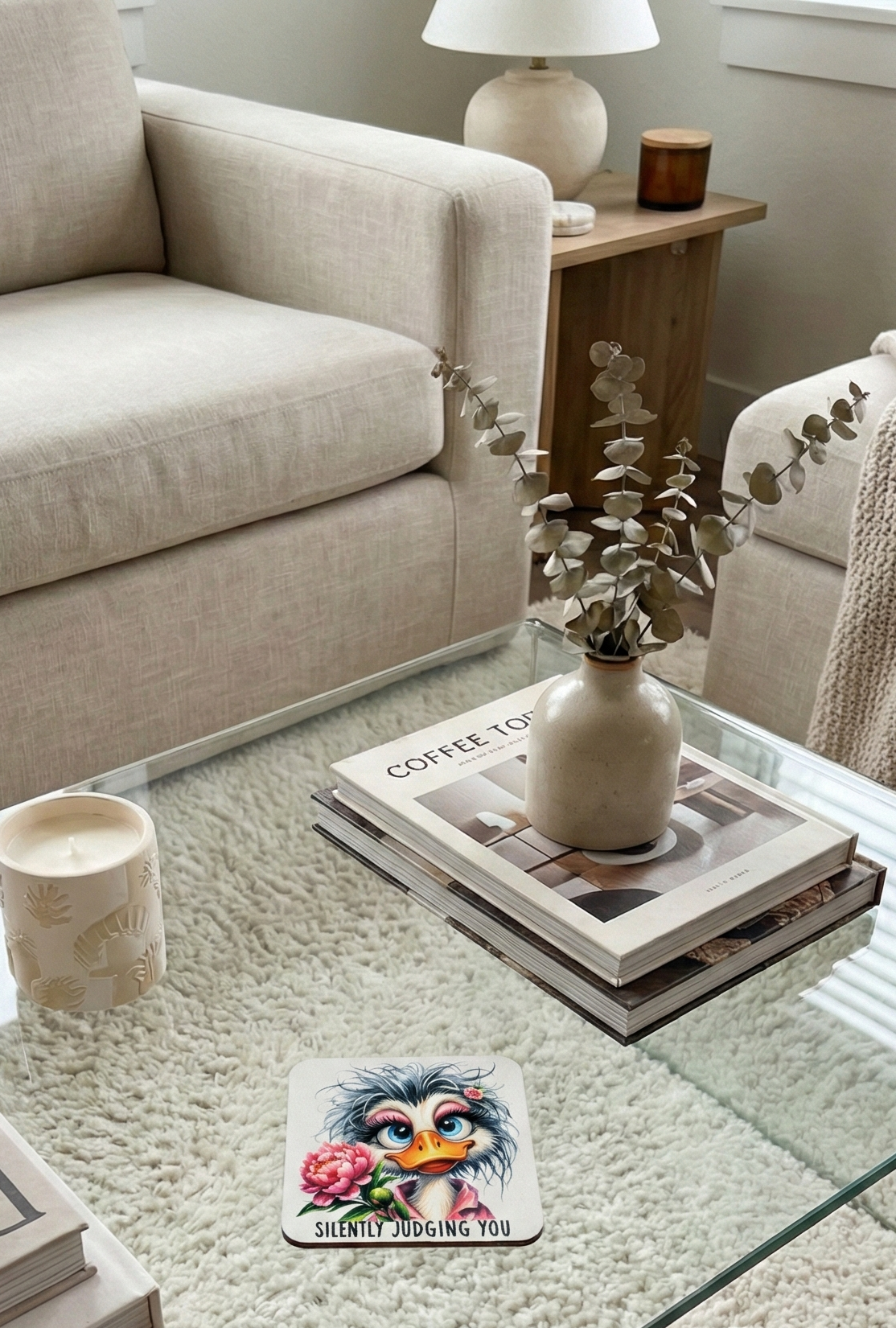 Light, neutral living room setting. A glass table holds books, an embossed candle, and a coaster showing a fluffy Grey duck holding a peony.