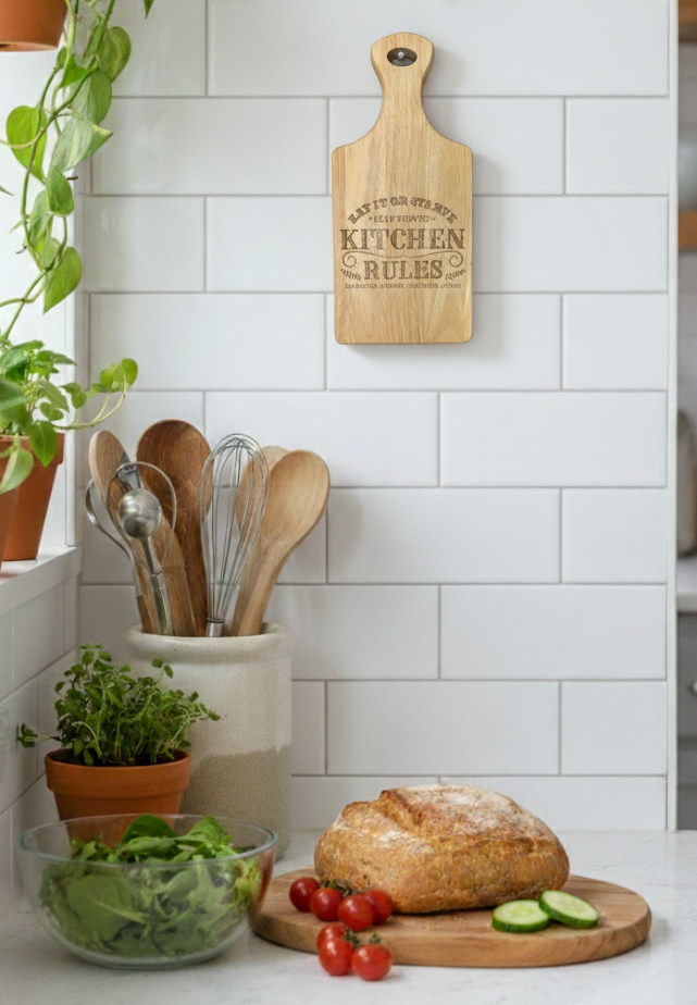 Light wooden cutting board hanging on a glossy white tiled wall. Rustic bread, green salad, and light wood utensils below.