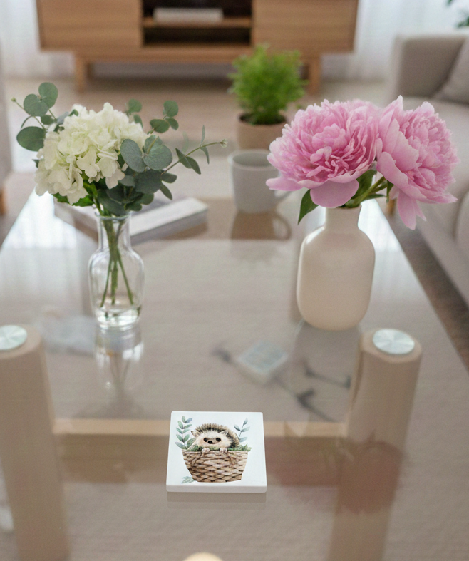 White square coaster on a glass table featuring a hedgehog illustration inside a brown woven basket with green leaves.