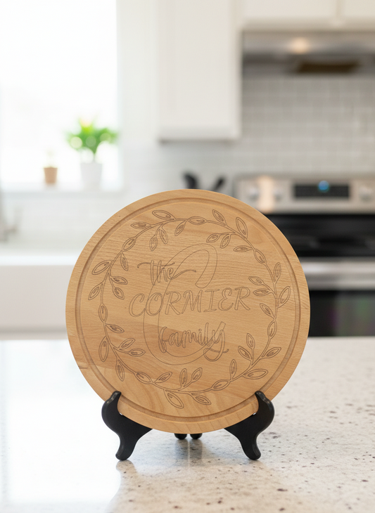 A round pale wood cutting board with detailed laser engraving of a wreath and text, displayed upright on a matt black stand.