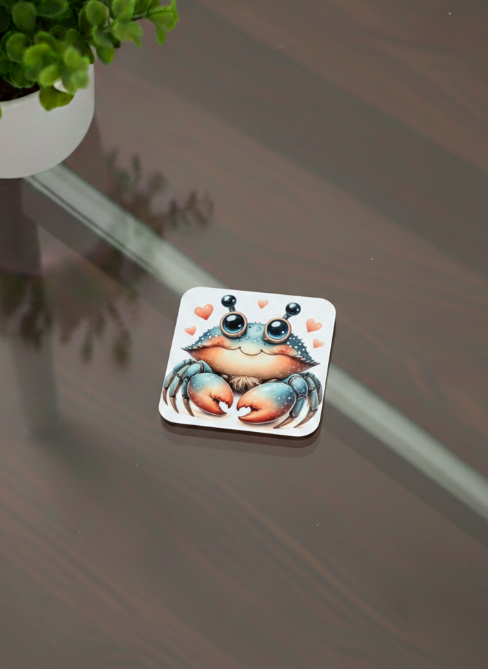 A square coaster with a cute blue and orange crab illustration, featuring large eyes and surrounded by pink hearts.