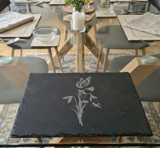 Dark grey slate placemat with an etched design of snowdrops and a butterfly, on a clear glass table.