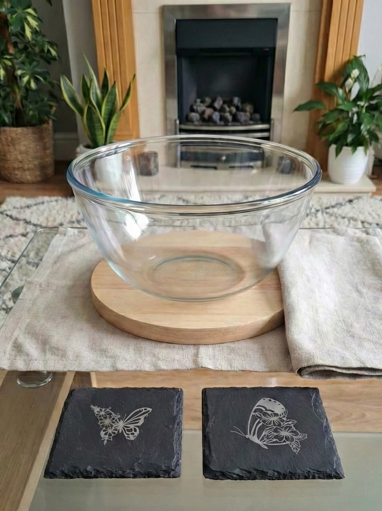 Two square slate coasters with butterfly engravings, displayed on a table with a fireplace in the background.