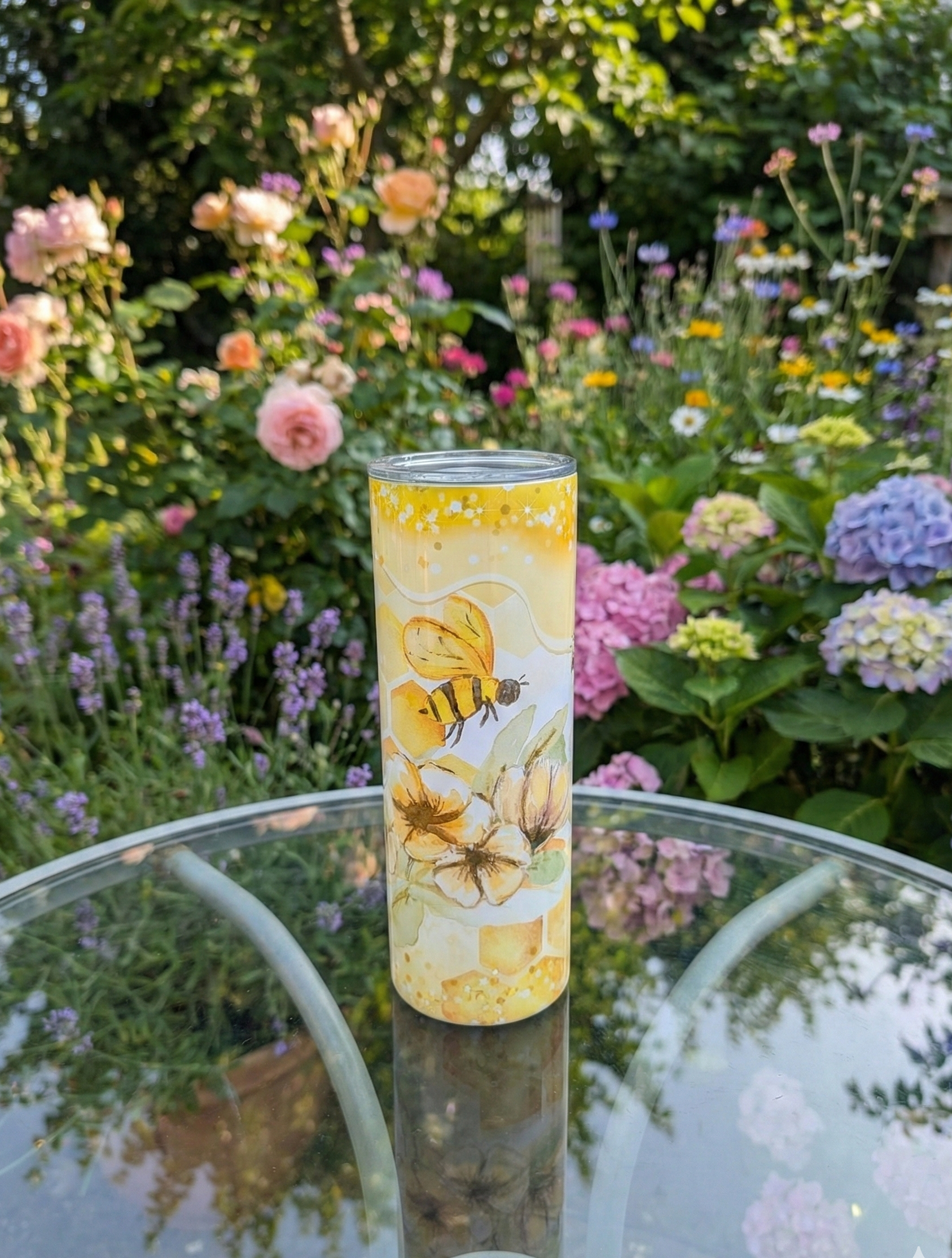 Slim tumbler with a honeybee illustration on a circular glass table, with a colourful, blooming garden in the background.