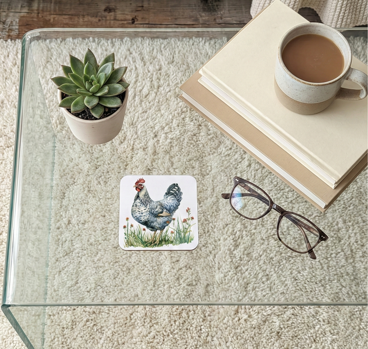 Close-up of a square coaster showing a speckled Grey chicken standing in a meadow illustration on a glass table.