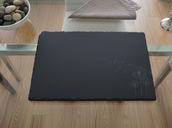 Dark grey slate placemat with textured edges and a white engraving of dandelion seeds blowing in the wind.