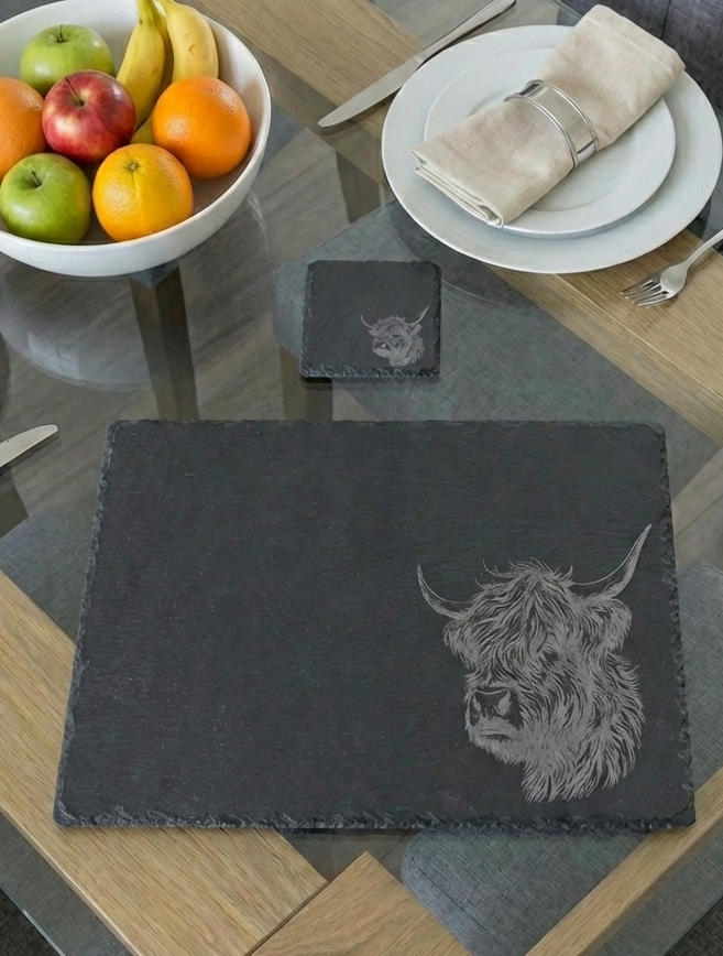 Dark grey slate placemat with a detailed Highland cow etching. White fruit bowl, plates and silver cutlery on glass table.