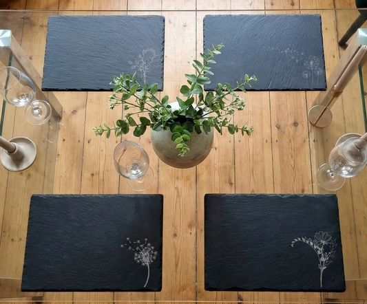 Top-down view of four charcoal grey slate placemats with white floral engravings on a glass-topped wooden table.
