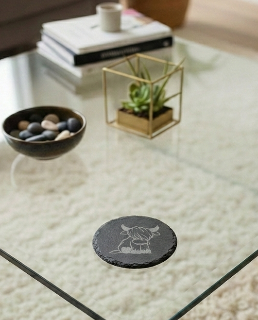 Round charcoal grey slate coaster featuring an etched profile of a Highland cow, resting on a glass surface.