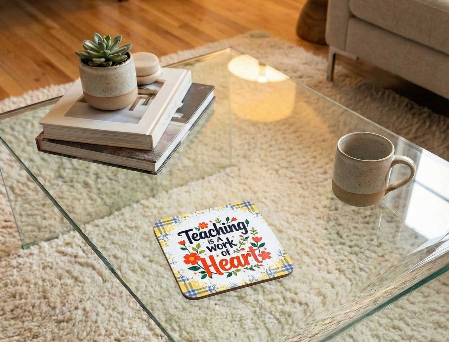 Yellow checked coaster with floral motifs and 'Teaching is a work of heart' on a glass table near a cream mug.