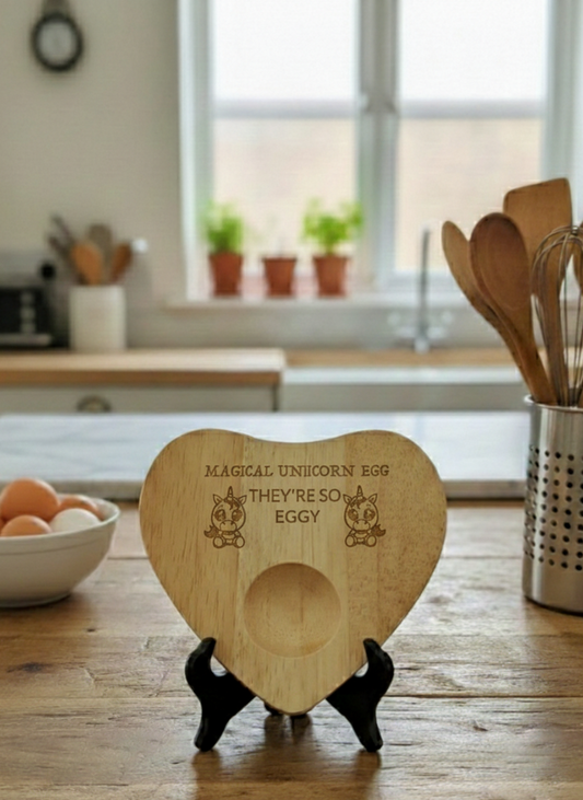 Heart-shaped light brown wooden egg stand, engraved with 'MAGICAL UNICORN EGG', on a rustic kitchen table.