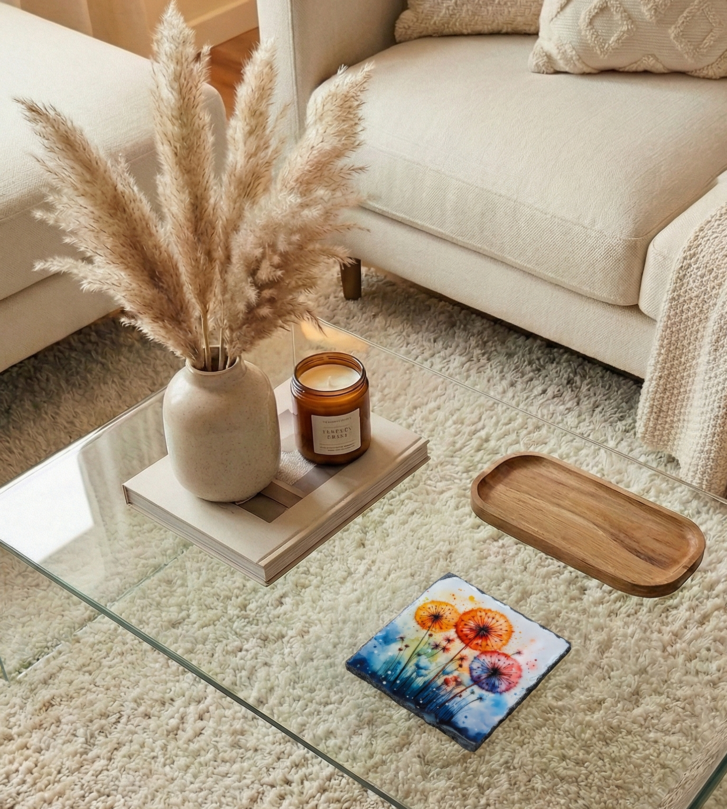 Colourful square coaster with orange and pink floral art sits on a glass table amongst soft neutral decor and textures.
