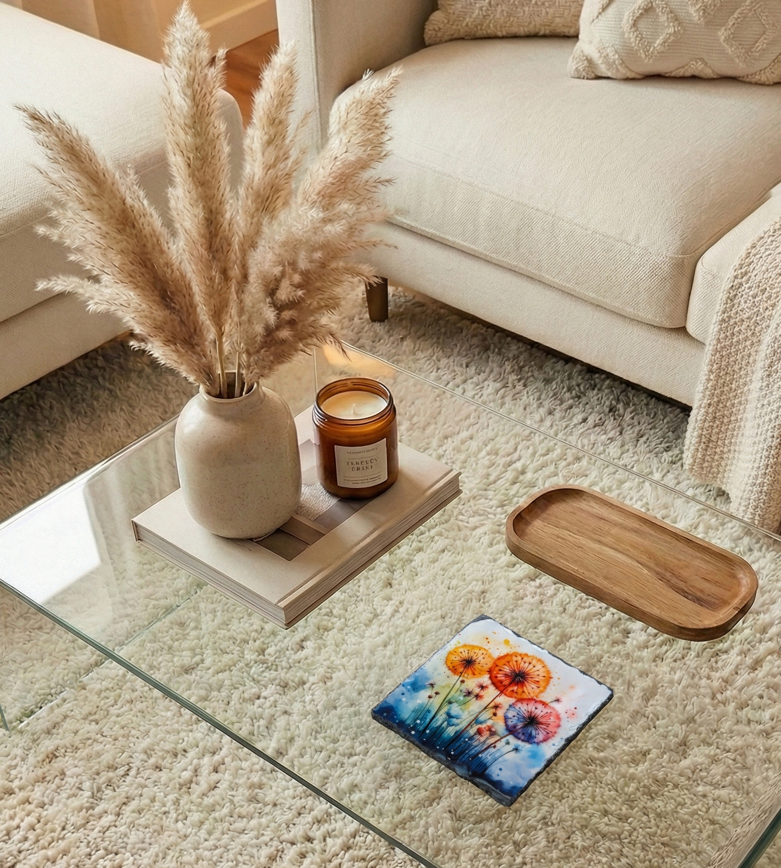 Colourful square coaster with orange and pink floral art sits on a glass table amongst soft neutral decor and textures.