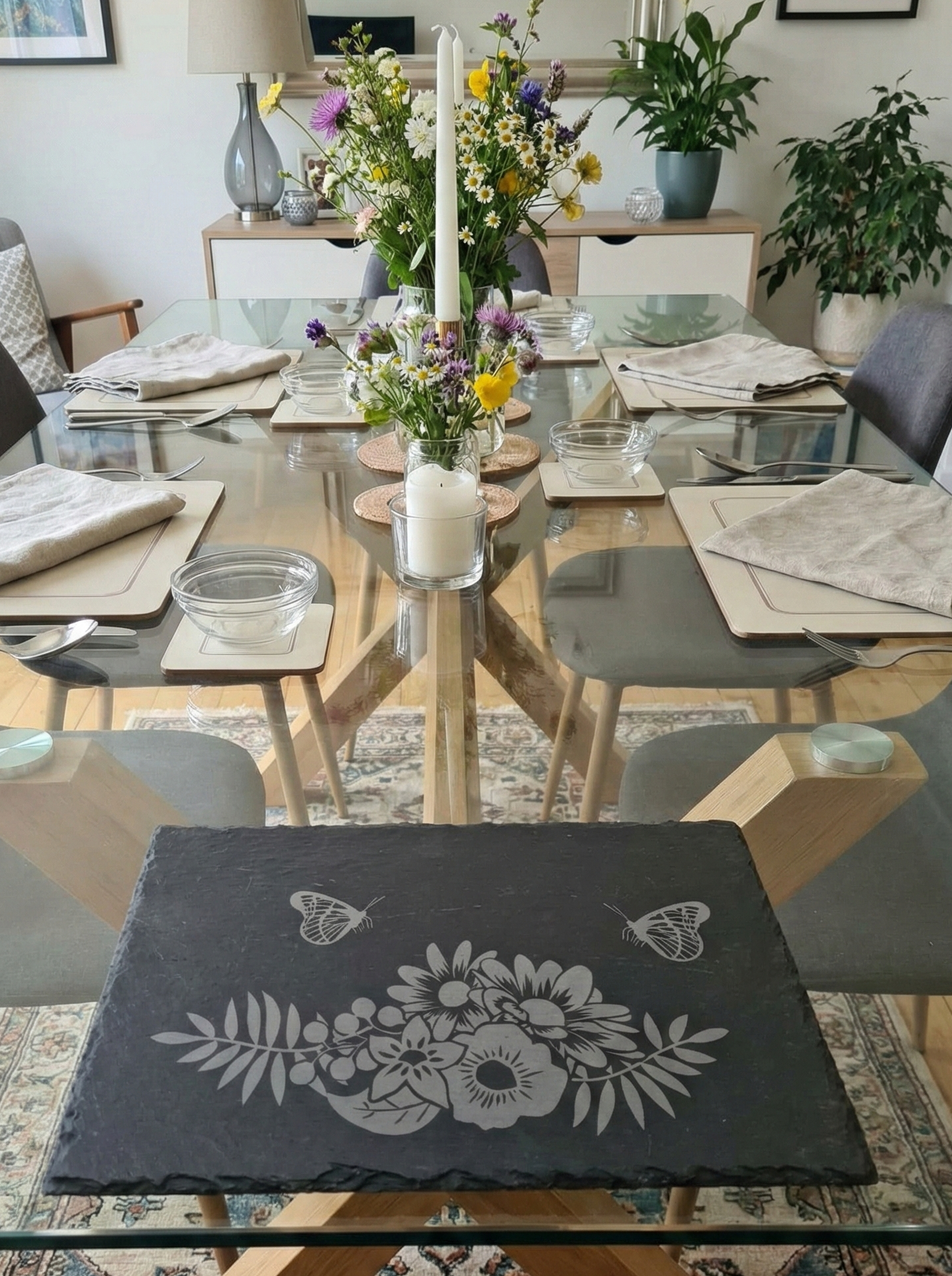 Rectangular slate placemat with grey floral and butterfly engravings on a glass table set for dinner.