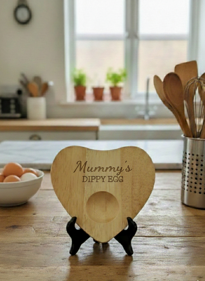 Heart-shaped blonde wooden dippy egg board, engraved "Mummy's DIPPY EGG," on a black stand. Bright kitchen setting.