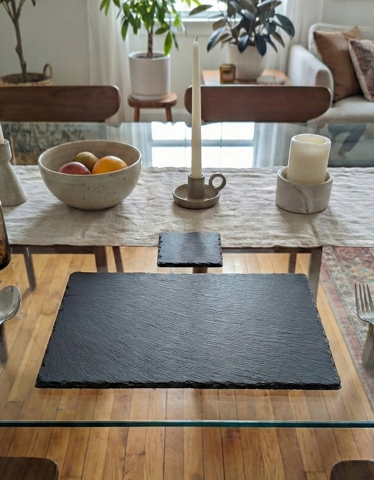 Rectangular charcoal-grey slate placemat and square coaster with rough textures on a glass dining table.