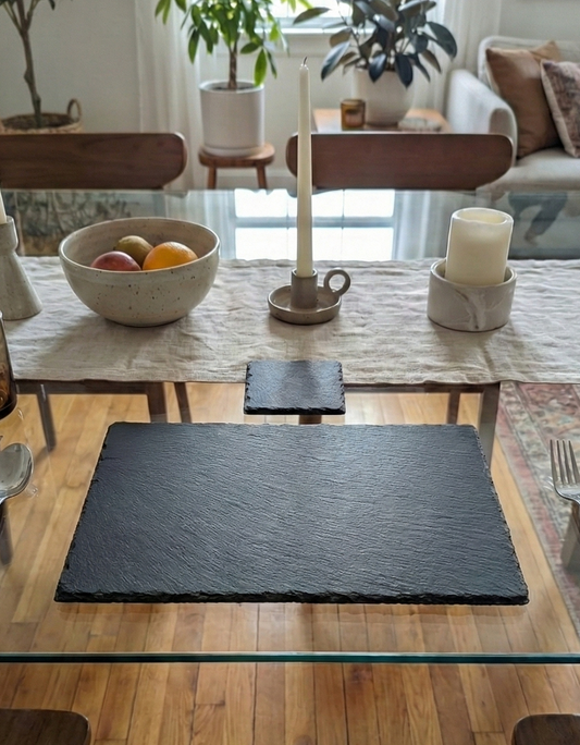 Rectangular charcoal-grey slate placemat and square coaster with rough textures on a glass dining table.