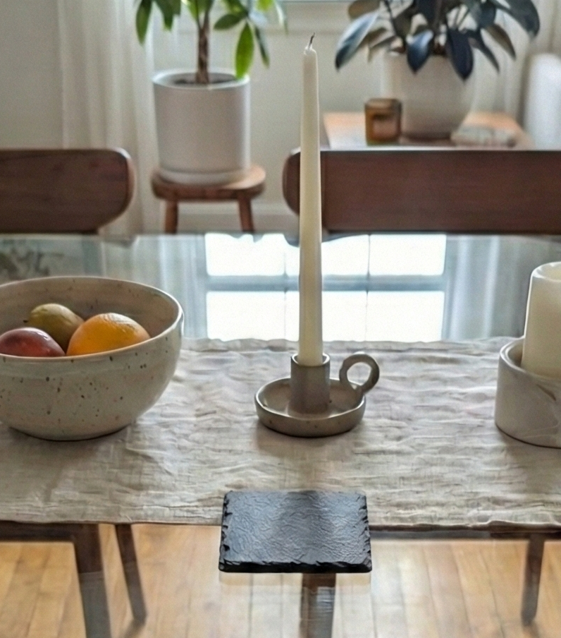 Close-up of a square, textured charcoal-grey slate coaster on a glass table with a bowl of fruit and candle.