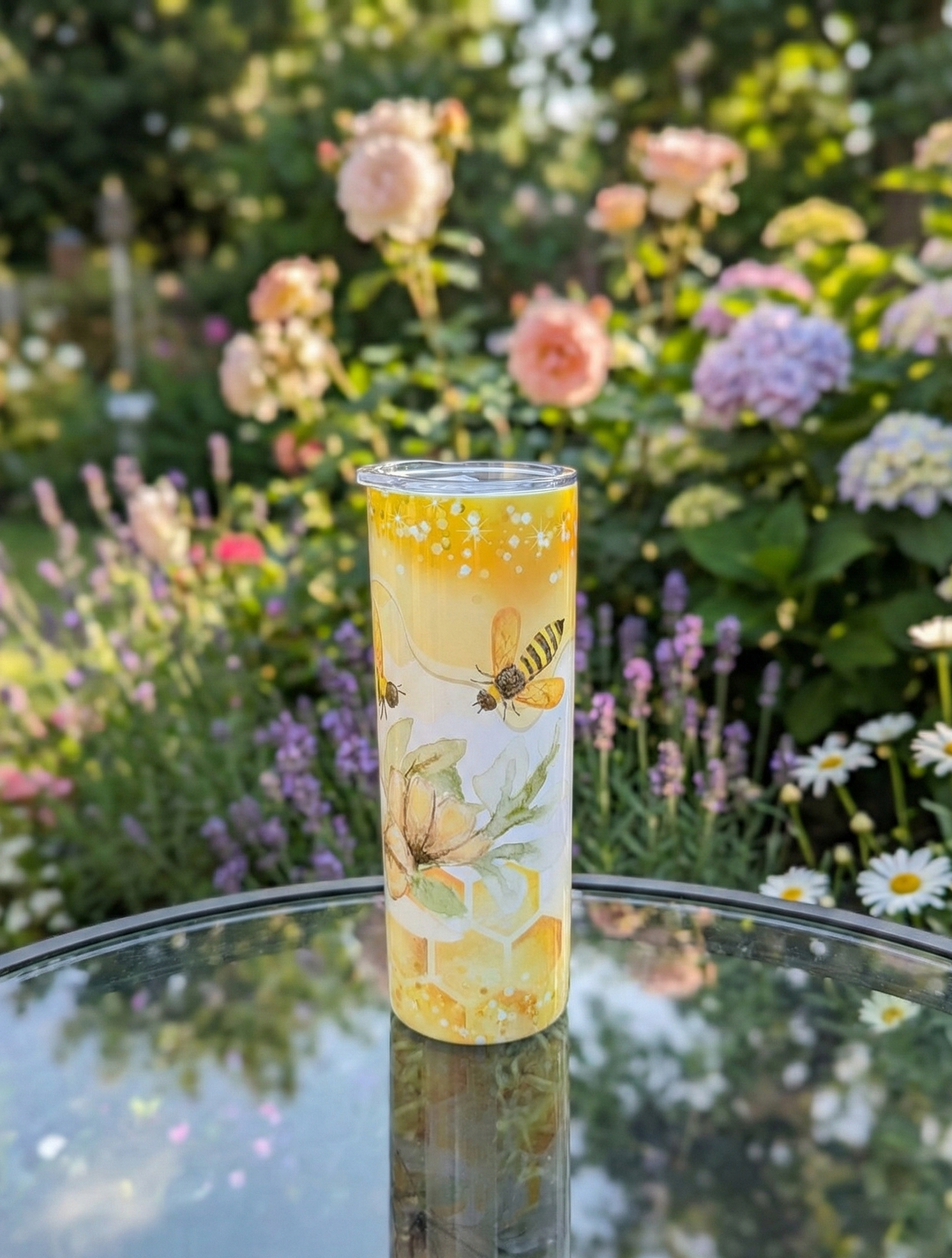 Yellow honeybee tumbler reflected on a glass table. The backdrop is a soft-focus garden with pink roses and purple flowers.