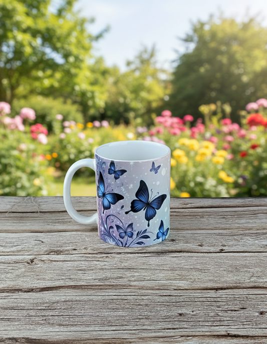 A white ceramic mug featuring a purple and blue butterfly design, sitting on a rustic wooden table in a sunny garden filled with blurred colorful flowers and trees.