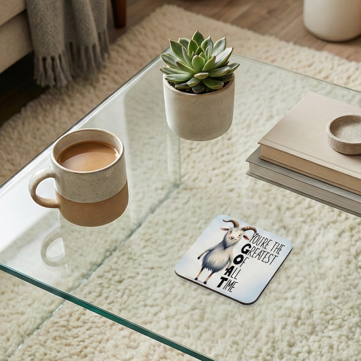Goat illustration on a square coaster beside a speckled ceramic mug and a green succulent on a glass table.