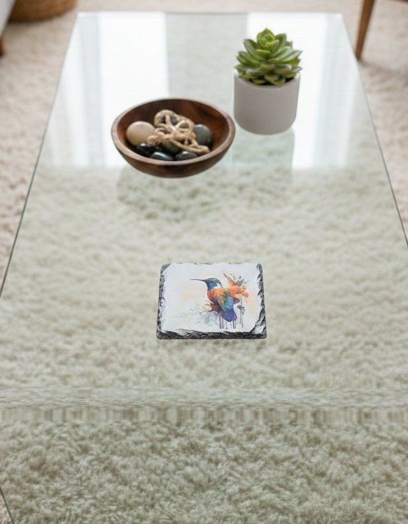 Single square slate coaster displaying a blue and orange hummingbird design. It sits on a glass table over a thick cream shag carpet.