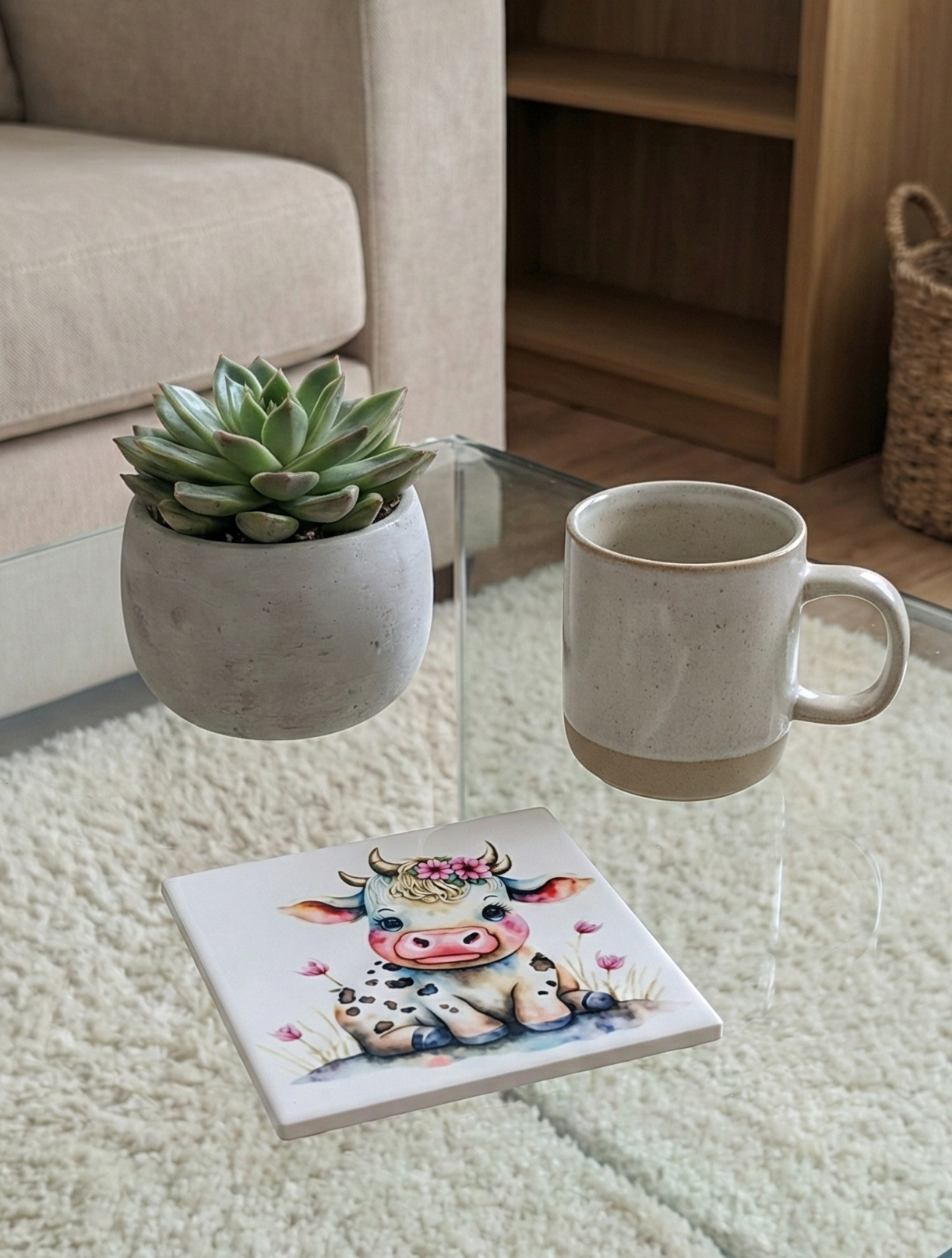 Watercolour cow coaster (black spots, pink flowers) on a glass table. A succulent and beige mug float above a thick, shaggy cream rug.