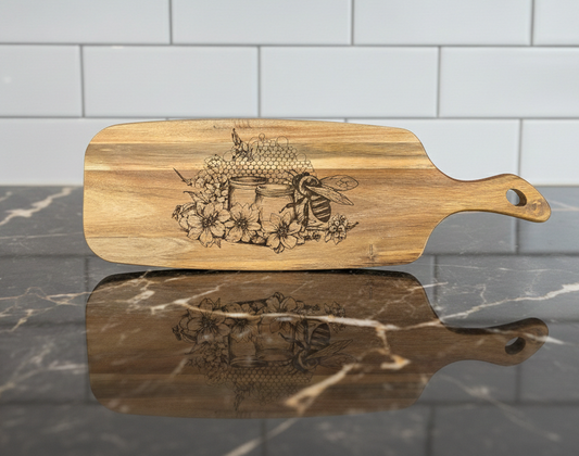 Richly grained wood cutting board, engraved with a detailed illustration of a bee, honeycomb, and florals. On a reflective dark counter.