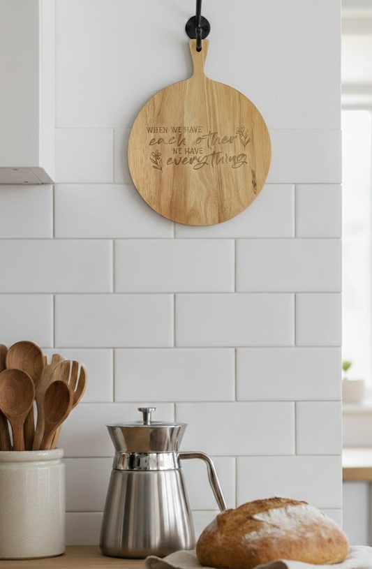 Light wood engraved serving board hangs on a bright white subway tiled wall above a polished stainless steel coffee pot and bread.
