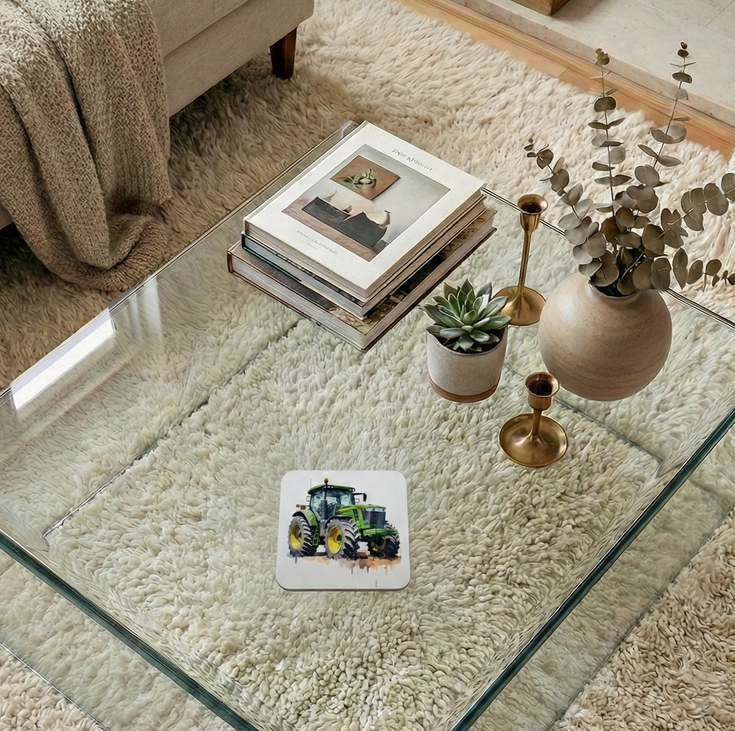 A white coaster with a green tractor illustration on a glass table, near a beige vase and a shaggy cream-coloured rug.