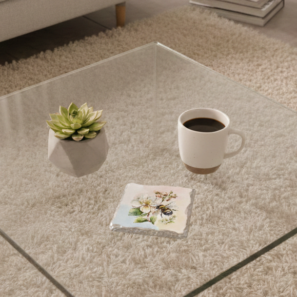 Stone coaster with a bee and white blossom watercolour on a glass table, next to a succulent and a white coffee mug.