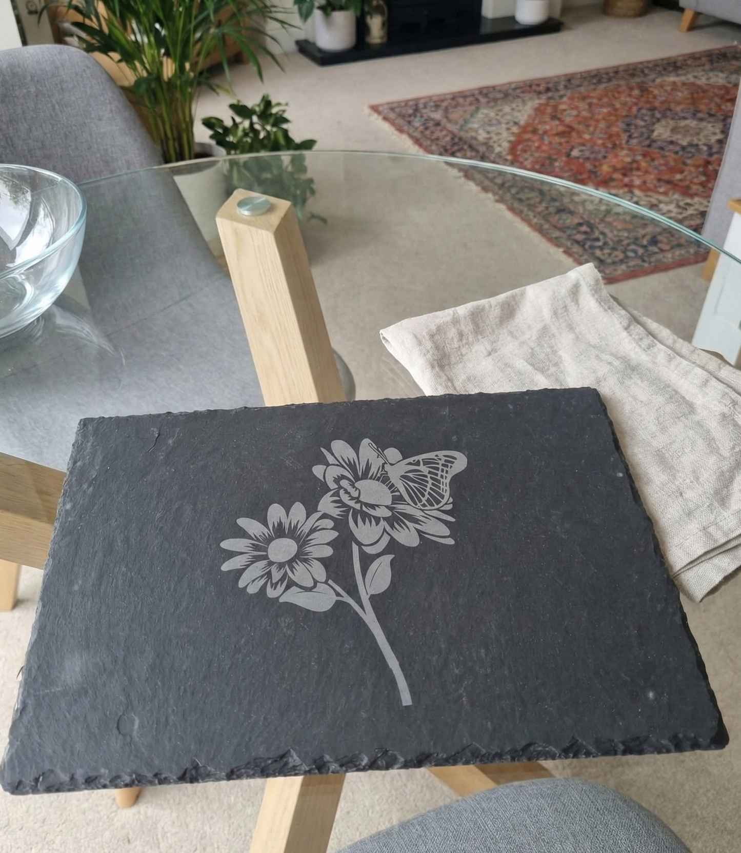 A dark grey slate placemat with an etched daisy and butterfly motif, placed on a glass-topped table.