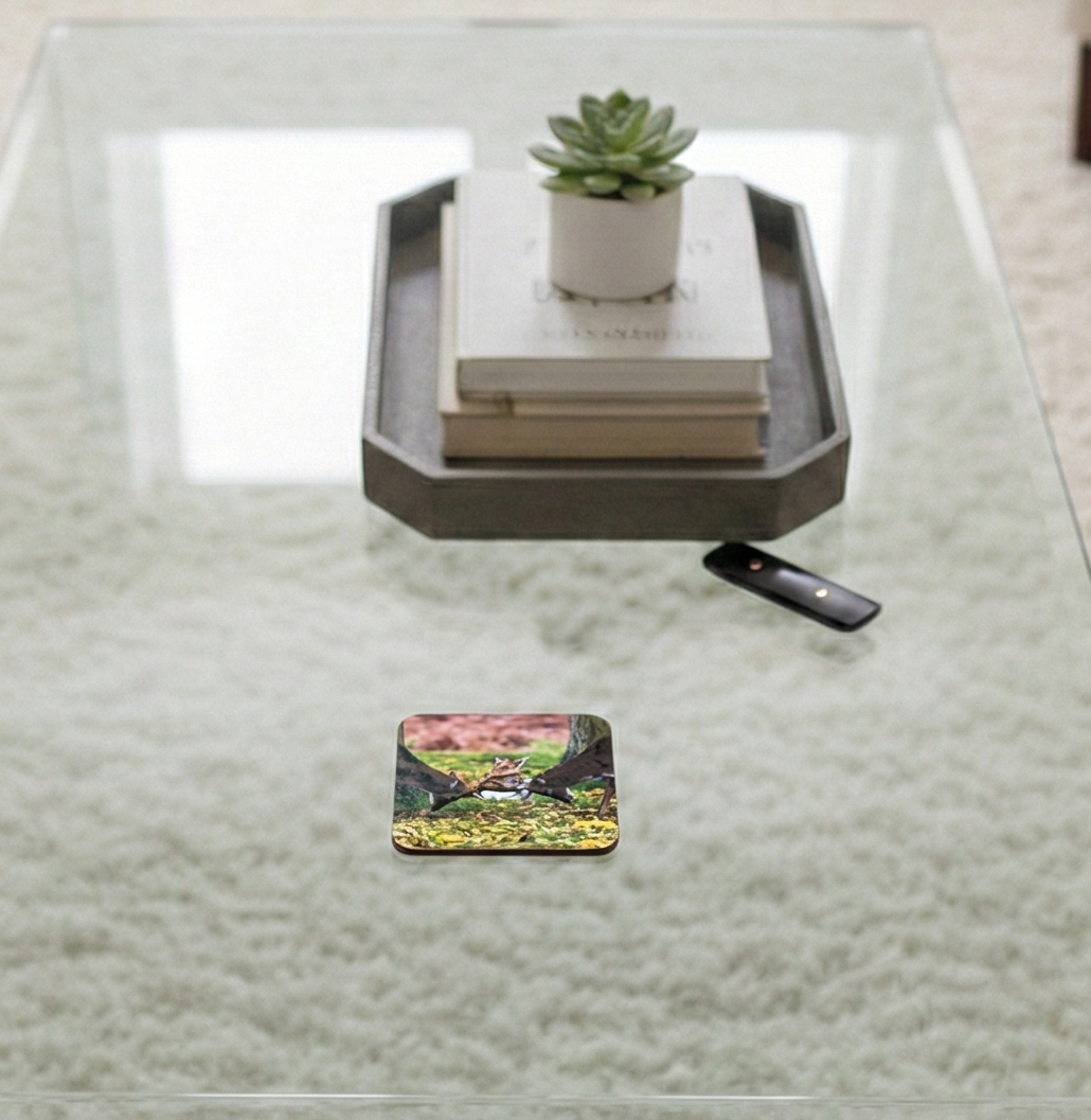 Square coaster showing two deer clashing on a glass table. A grey tray with a book and succulent is in the background.