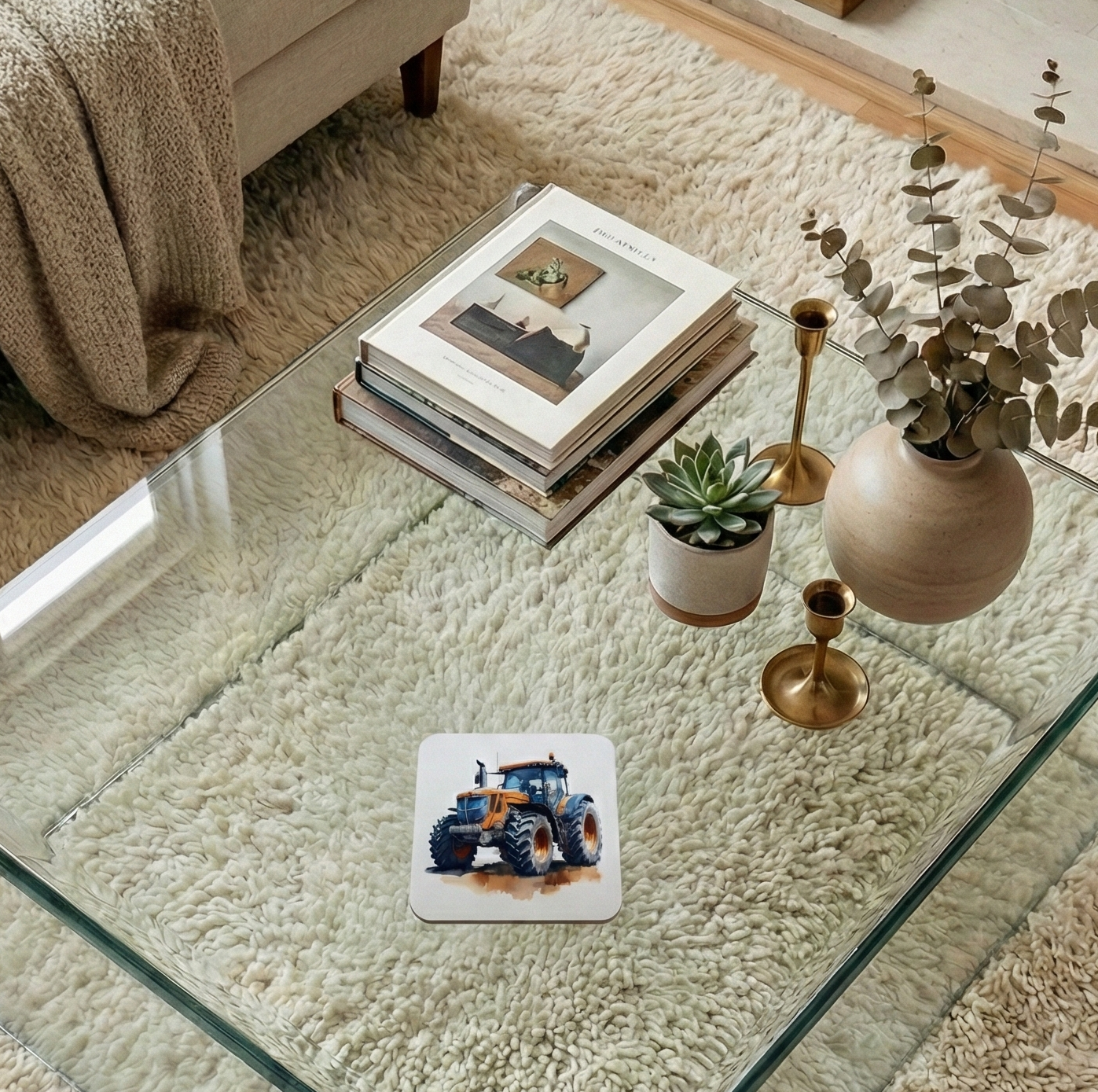 A square coaster featuring an orange tractor on a glass table, alongside a stack of books and gold candlesticks.