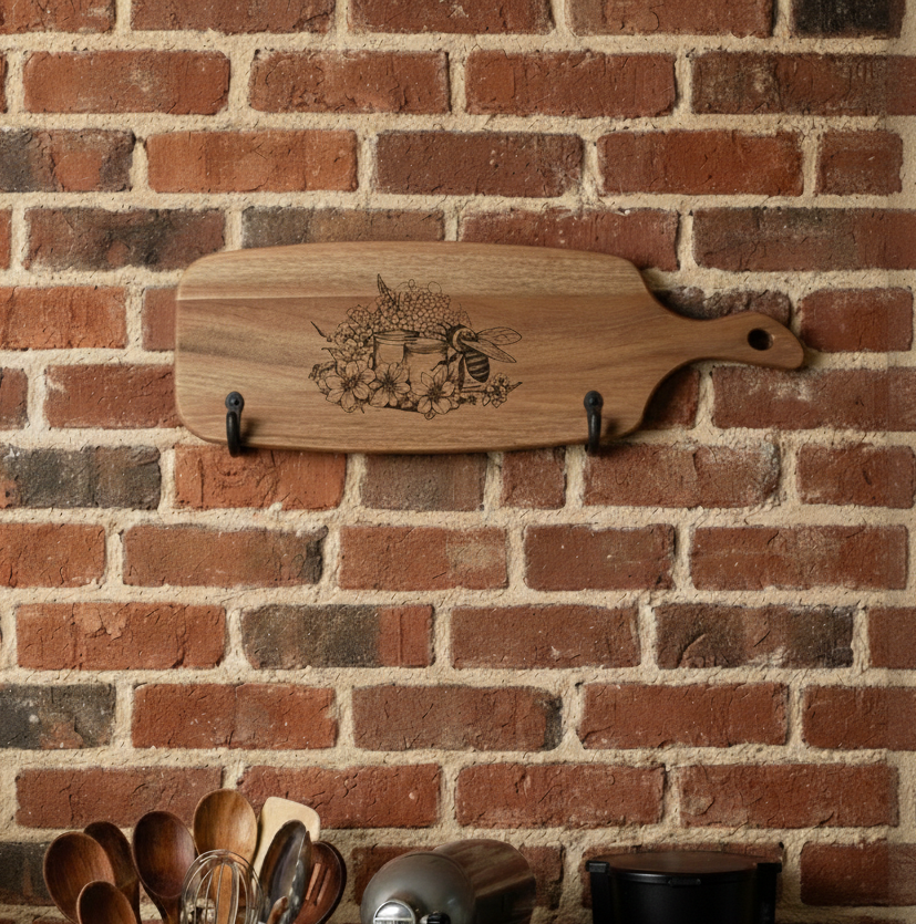Grained wooden cutting board, engraved with a bee and flowers, hanging on black metal hooks against a rustic red-brown brick wall.