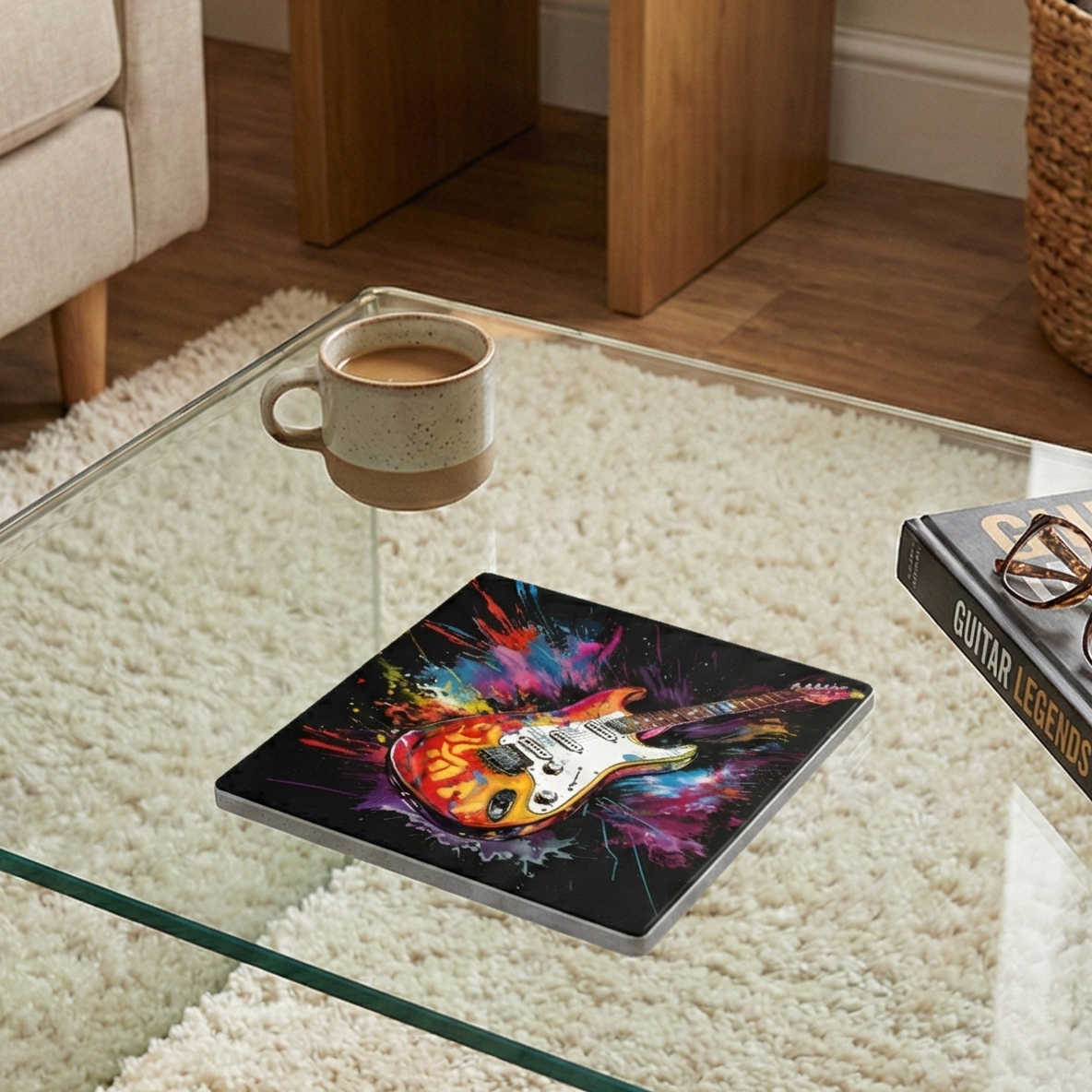 A square coaster with a colourful electric guitar and paint splatters on a glass table near a mug and a book.