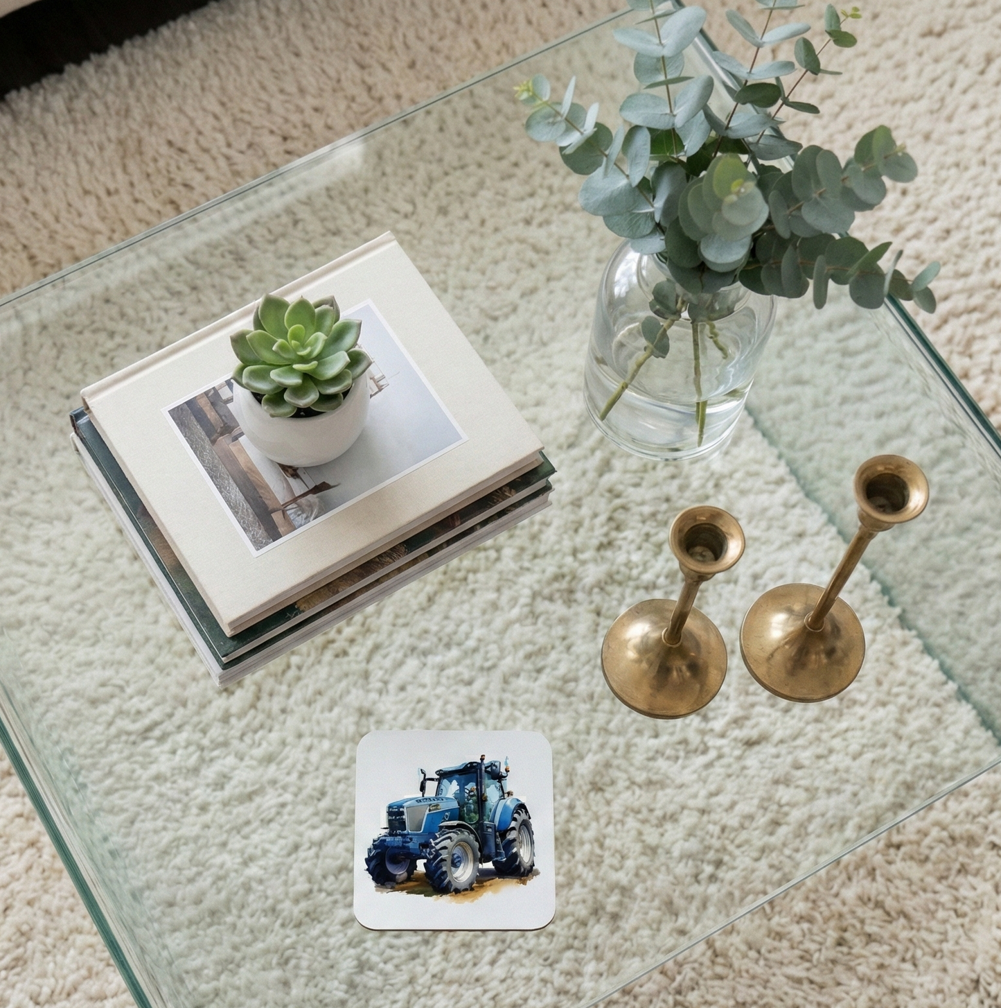 A white coaster with a blue tractor illustration on a glass table, with a succulent and gold candlesticks nearby.