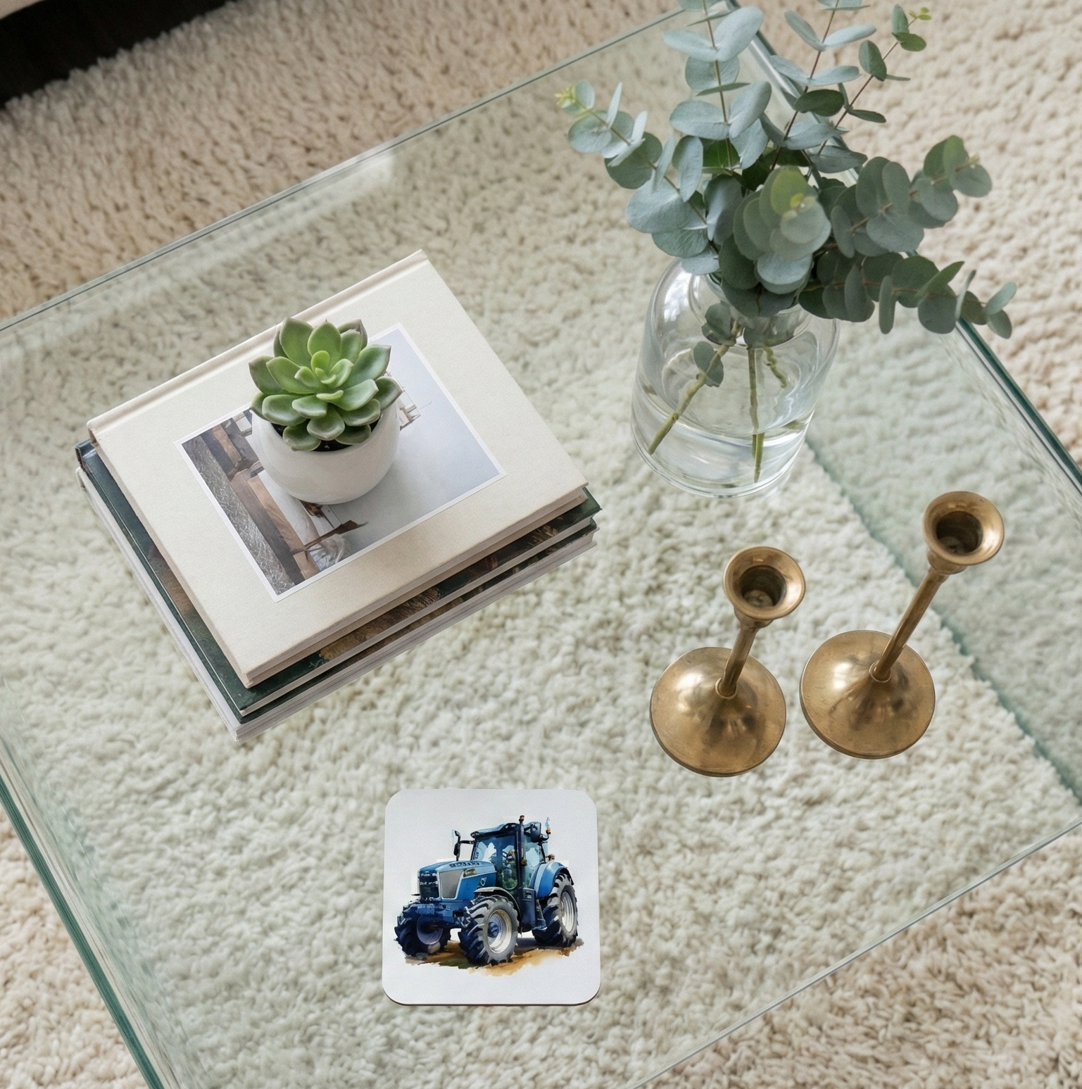 A white coaster with a blue tractor illustration on a glass table, with a succulent and gold candlesticks nearby.