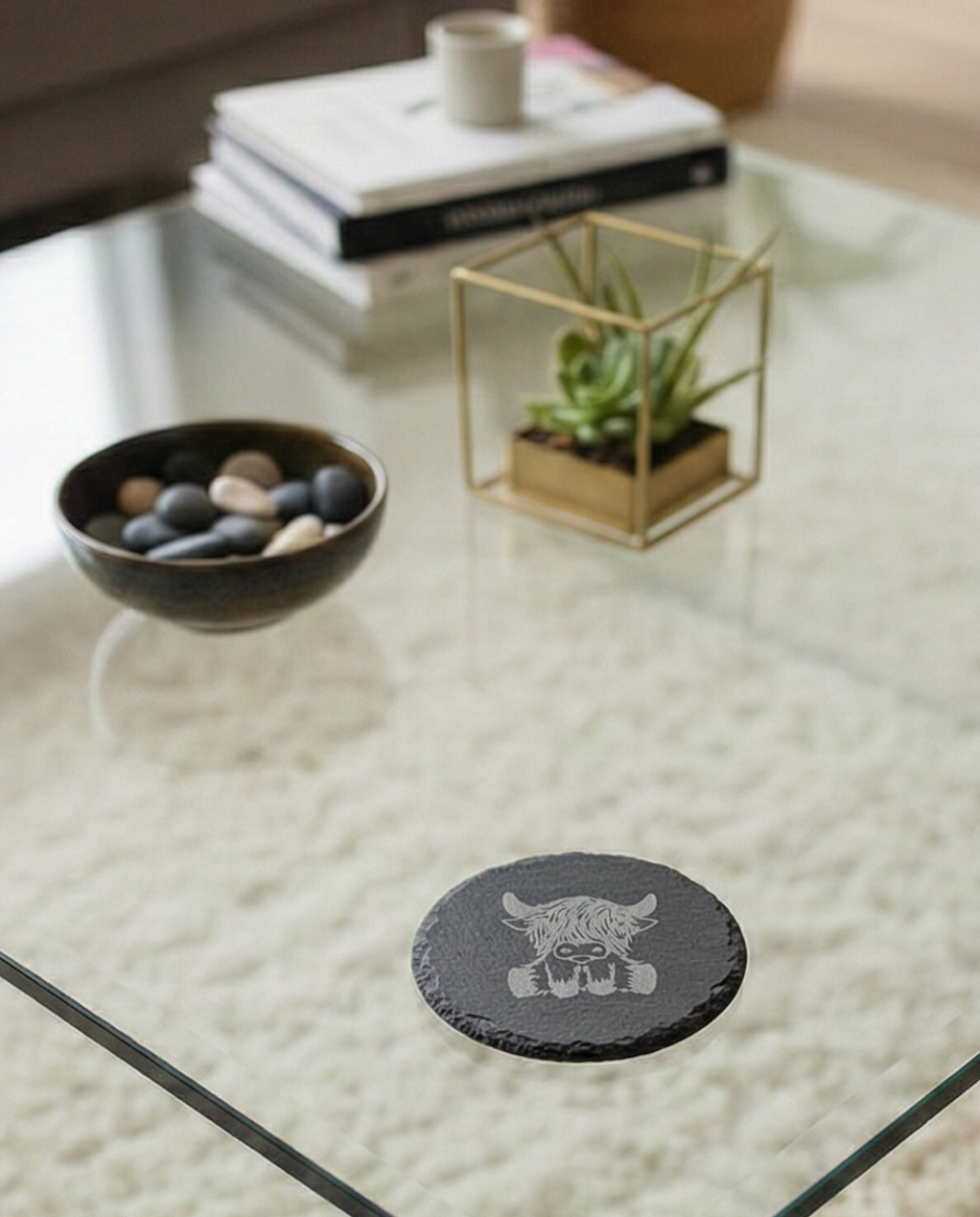 Circular grey slate coaster with an etched front-facing Highland cow, placed on a clear glass table.