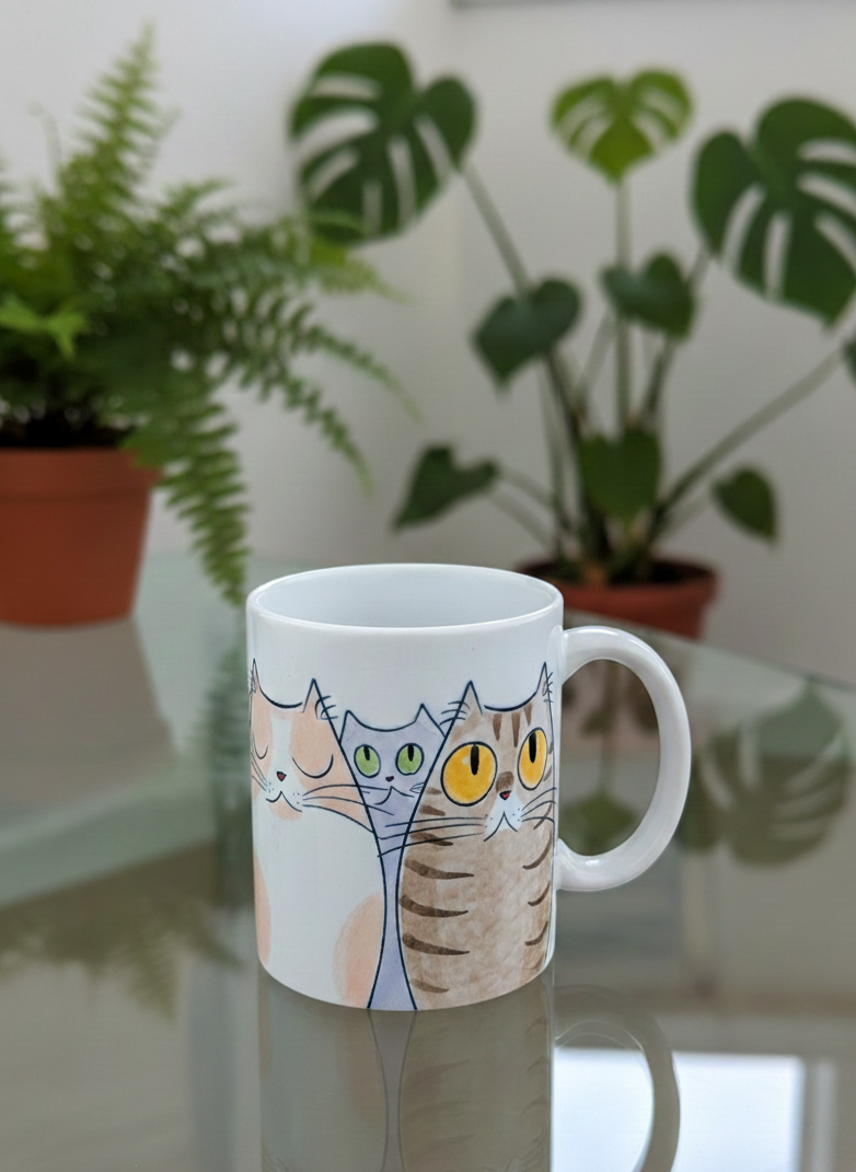 White ceramic mug showing cartoon cats in peach, grey, and brown stripes, with bright green and amber eyes.