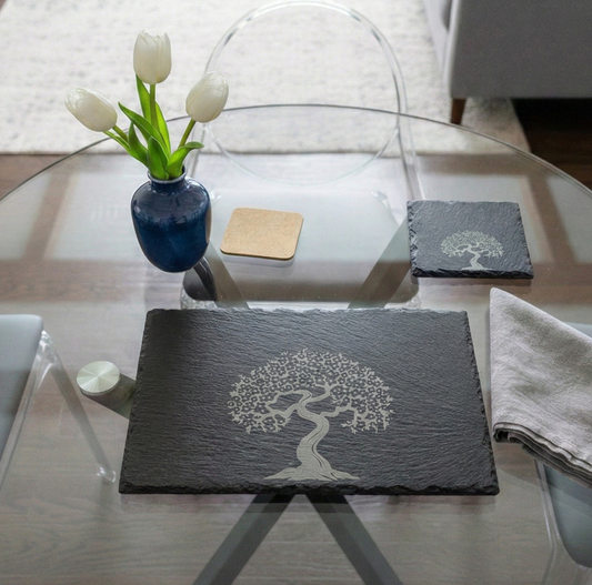 Rectangular black slate placemat with a grey tree engraving in the centre on a glass table with white tulips.