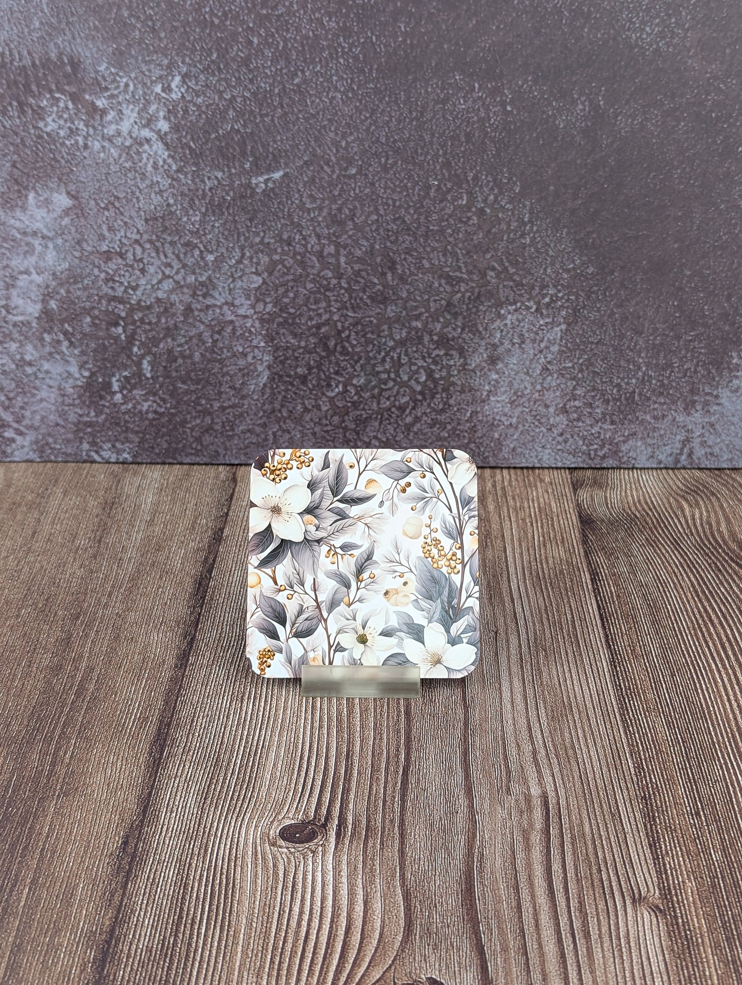 Close-up of a square coaster with grey and white botanical print, on a wooden surface with a textured grey background.